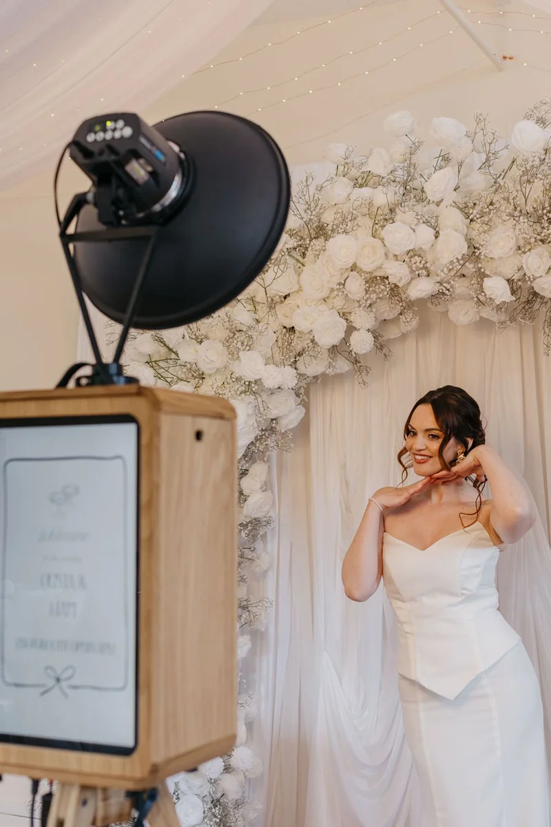 The oak Icon Booth beside a bride, with ivory drapes and white wedding flowers.